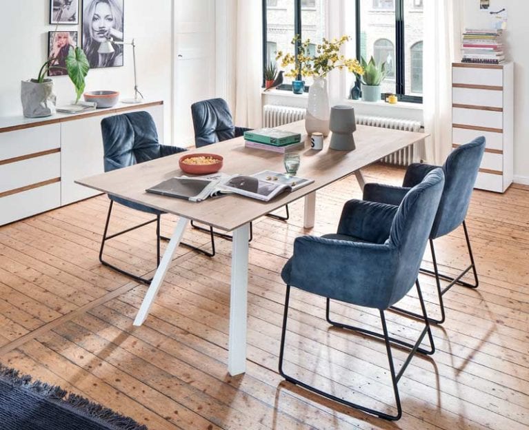 Foto eines Esstischs in Holzoptik mit weißen Beinen und vier bequemen Sesseln in blauer Samtoptik in einem hellen Essbereich