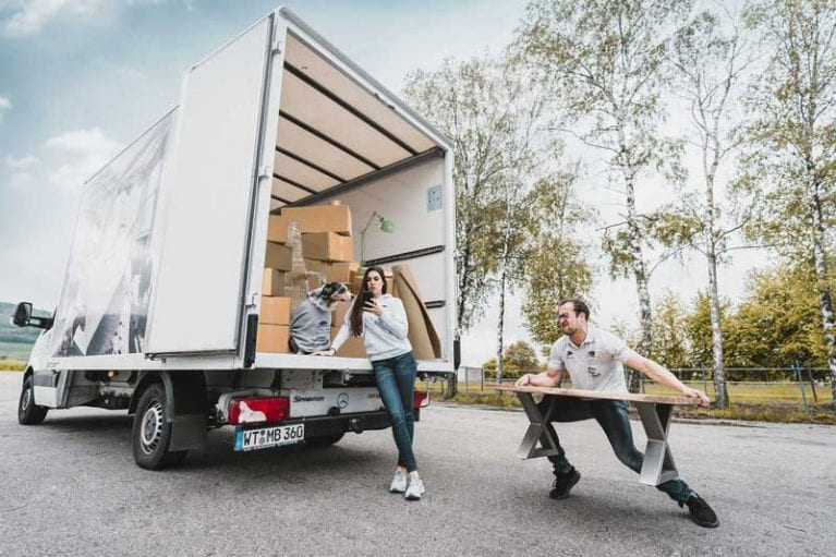 Foto von Inhaber Chris Beck, der einen Couchtisch in einen Transporter laden will - Freundin lehnt gelassen an Transporter, Hund sitzt in Transporter und beobachtet sein Herrchen