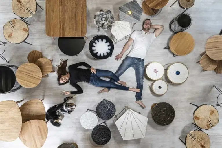 Foto von Inhaber Beck mit Freundin und Hund am Boden liegend, Perspektive von oben - um sie herum verschiedene Couchtischchen aus Holz und Marmor