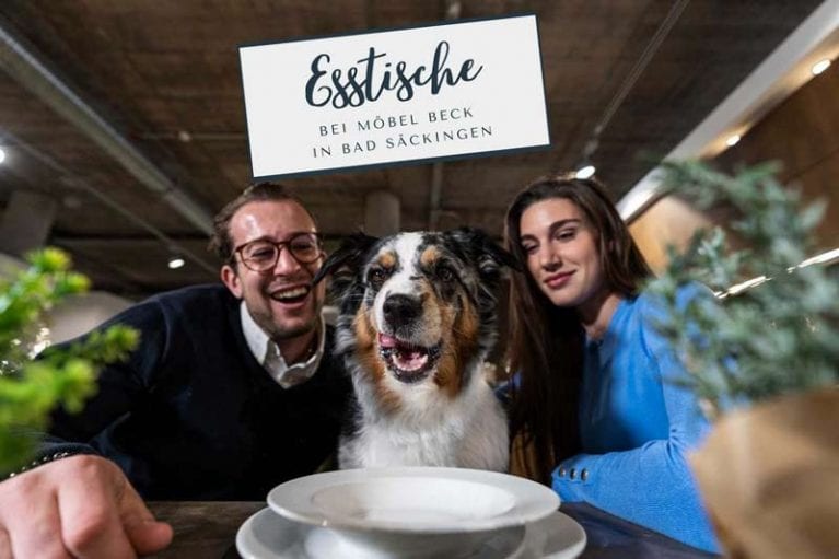 Christopher und Ines sitzen am Esstisch mit Ihrem Hund in der Mitte und einem Teller davor