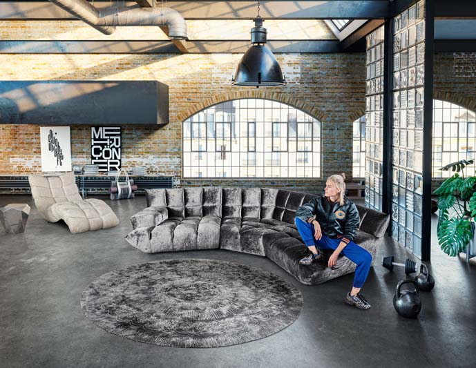 Graues samtiges Bretz Sofa Cloud 7 steht in einer Loftwohnung mit einer sportlich gekleideten Blondine auf dem Sofa.