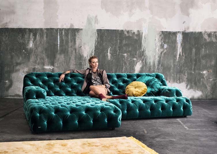Samtgrünes Bretz Sofa Cocoa Island mit schönem Steppmuster steht in einer industriellen Wohnung mit einer sehr schicken Dame darauf.
