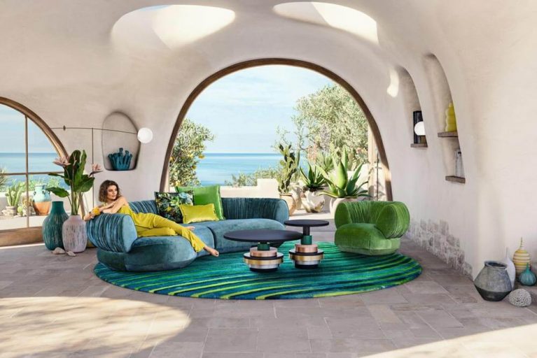 Bretz Designersofa NAMAMI in blau mit einer Dame in einem Senfgelben Jumpsuit darauf und einem grünen Sessel daneben.